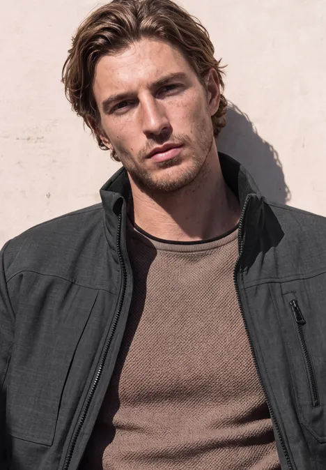 Veste à col montant et fermeture zip ash grey melange Ein junger Mann mit braunen Haaren in einem dunklen Jacket lehnt entspannt an einer hellen Wand.