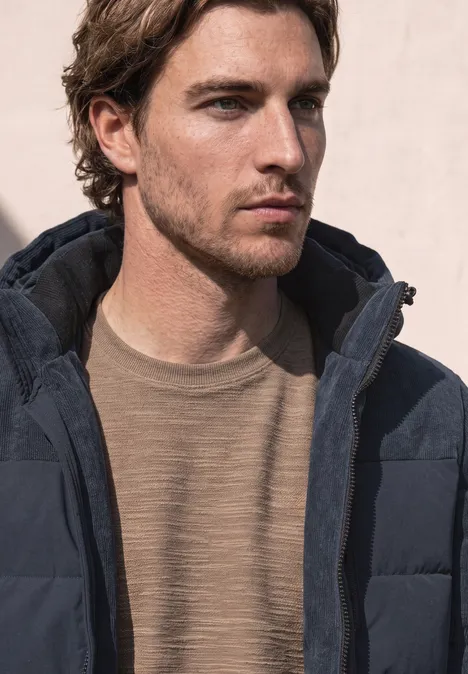 Ein Mann in einer dunklen Steppjacke und beigem Pullover blickt nach rechts vor hellem Hintergrund.