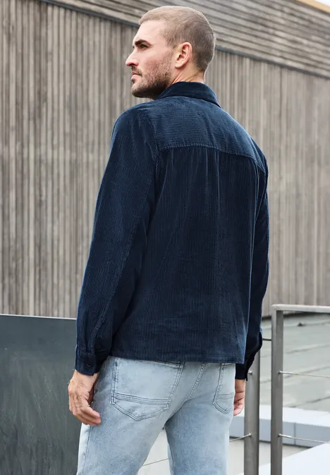 Un homme porte une chemise en velours côtelé bleu marine et un jean clair, debout sur une terrasse urbaine.