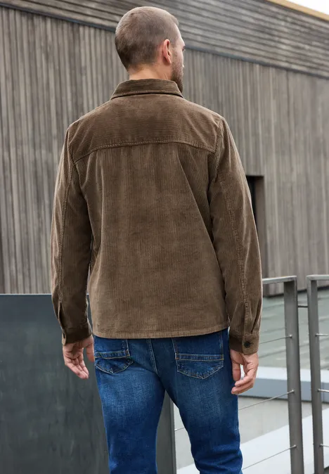 Homme de dos portant une veste en velours côtelé marron et un jean bleu, sur un toit moderne.
