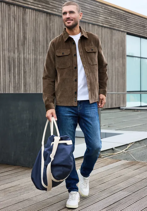 Un homme en veste marron et jean sur une terrasse en bois, tenant un sac de voyage bleu.