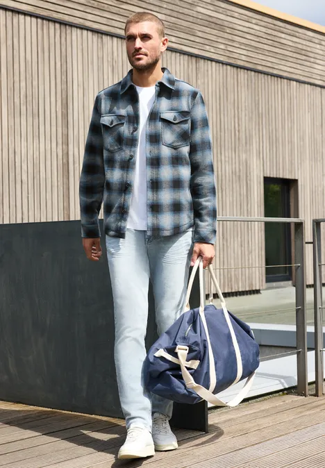 Un homme en chemise à carreaux et jean porte un sac de voyage sur une terrasse devant une maison en bois.