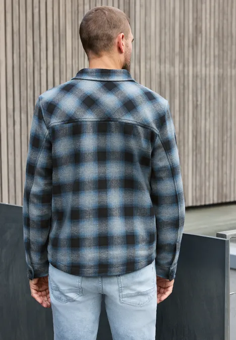 Un homme vu de dos porte une chemise à carreaux bleu-noir et un jean clair devant un mur de bois.