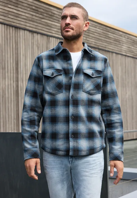 Un homme porte une chemise à carreaux bleu-gris, un T-shirt blanc et un jean clair devant un mur en bois.