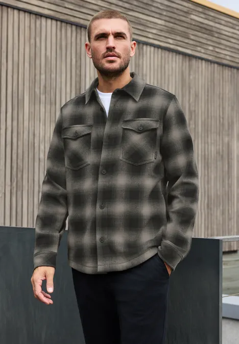 Un homme porte une chemise à carreaux vert et noir devant un mur en bois.