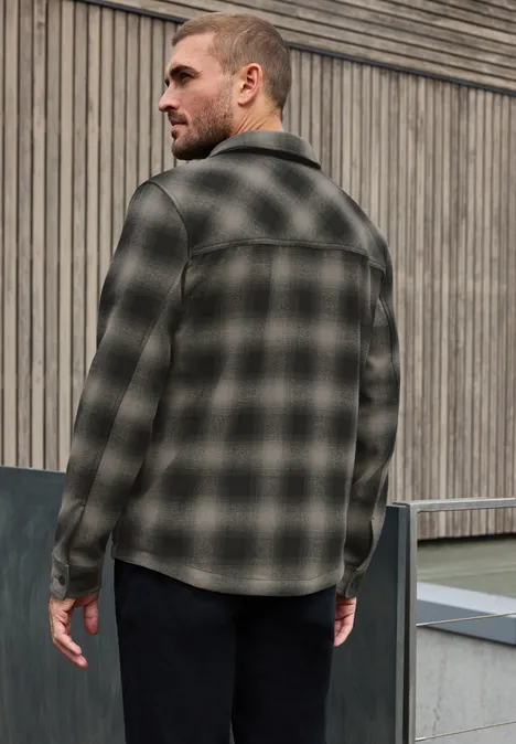 Homme de dos en chemise à carreaux sobre et pantalon sombre devant une façade en bois.