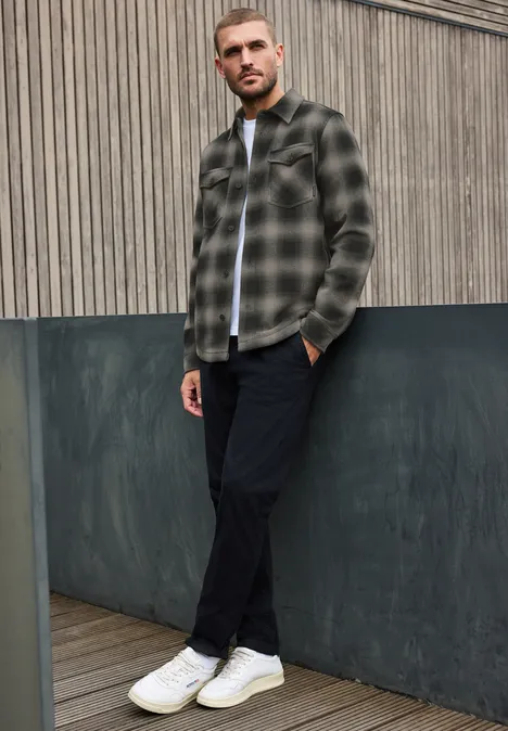 Un homme appuyé devant une façade grise, portant une chemise à carreaux, un pantalon noir et des baskets blanches