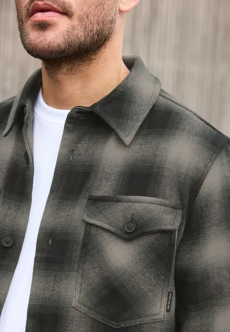 Gros plan d'un homme en surchemise à carreaux vert foncé et noir avec poches poitrine, sur un t-shirt blanc.