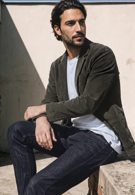 Ein Mann in Wildlederjacke, T-Shirt und Jeans sitzt entspannt auf einer Betonmauer im warmen Sonnenschein.