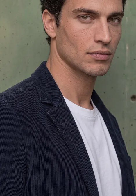 Junger Mann mit dunklem Blazer und weißem T-Shirt vor grünem Hintergrund, ernster Blick.