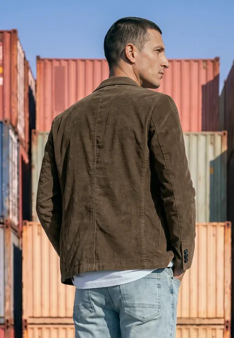 Ein Mann in brauner Jacke und Jeans steht seitlich vor bunten Containern.