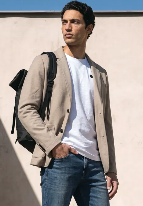 Ein Mann steht vor einer Wand, trägt eine beige Jacke, weißes T-Shirt, blaue Jeans und einen Rucksack.