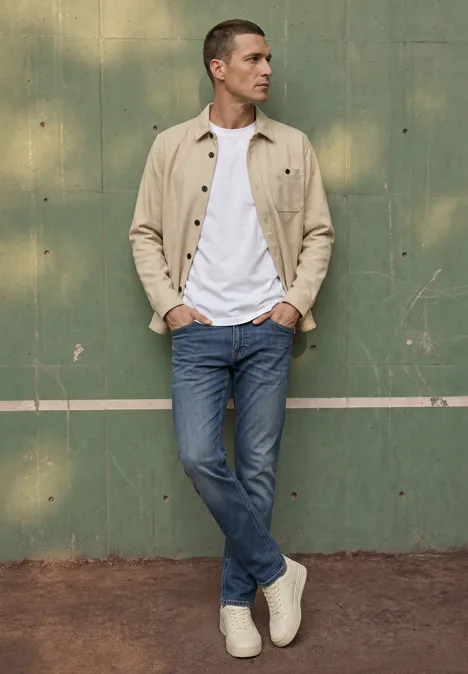 Ein Mann lehnt in beiger Jacke, weißem T-Shirt und Jeans vor einer grünen Wand mit weißem Streifen.
