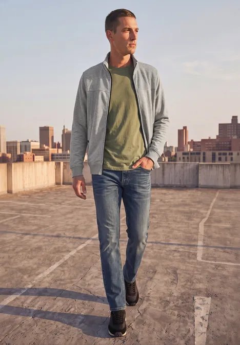 Ein Mann in Jeans, grünem Shirt und grauer Jacke läuft über ein Parkdeck mit Stadtkulisse im Hintergrund.