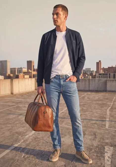 Ein Mann in Jeans, weißem T-Shirt und leichter Jacke mit Lederreisetasche auf einem Parkdeck vor der Stadt.