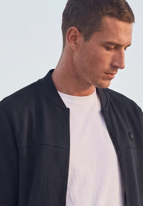 Ein junger Mann trägt ein weißes T-Shirt und eine dunkle Jacke und schaut nach unten.