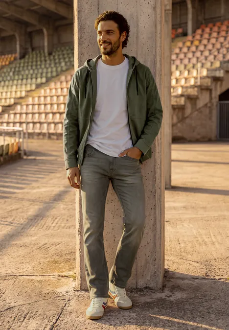 Ein lächelnder Mann in Jeans und Jacke lehnt an einer Betonstütze im Stadion.