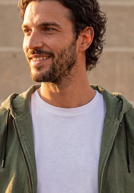 Ein lächelnder Mann mit Bart trägt ein weißes T-Shirt und eine grüne Kapuzenjacke im Freien.