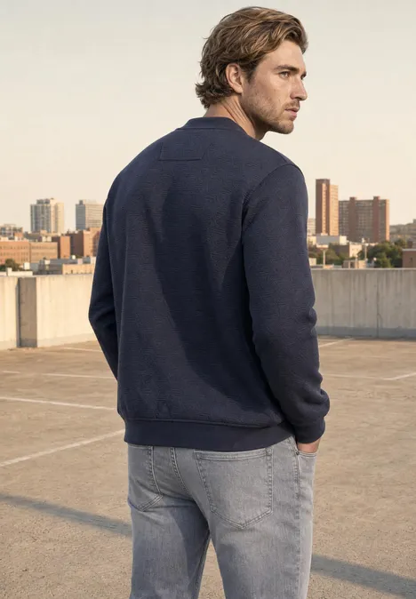 Blouson à structure 3D et fermeture zip dark ink blue Ein Mann steht mit dem Rücken zur Kamera auf einem Parkhausdach, trägt Pullover und Jeans, Stadt im Hintergrund.