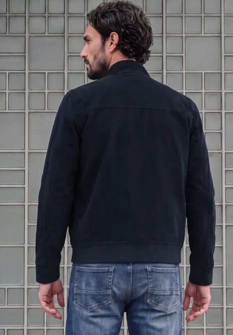 Ein Mann steht mit dem Rücken zur Kamera, trägt eine schwarze Jacke und Jeans vor grauer Wand.