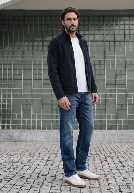 Ein Mann in Jeans, weißem T-Shirt und dunklem Cardigan steht vor einer grünen Fliesenwand auf Kopfsteinpflaster.