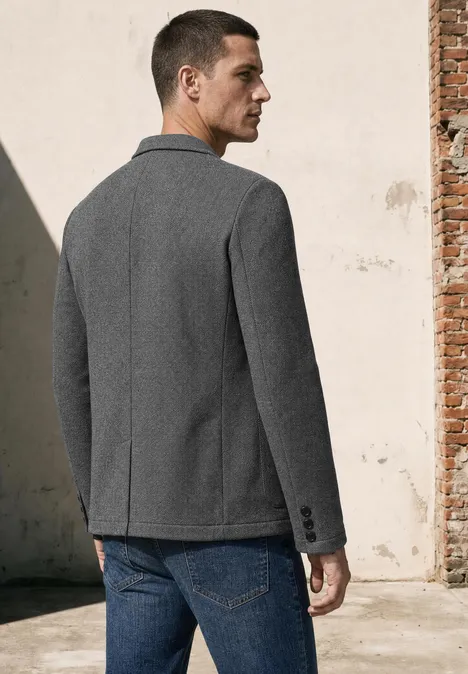 Ein Mann steht von hinten, trägt grauen Blazer und blaue Jeans vor einer hellen Wand.