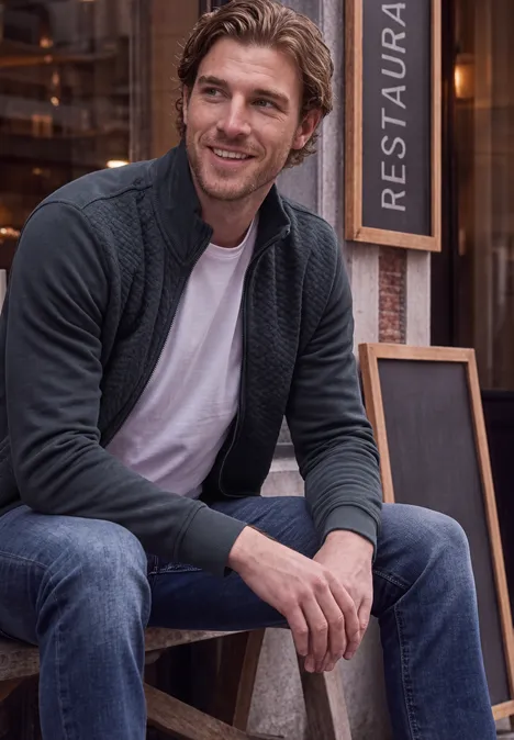 Un homme souriant assis devant un restaurant, en jean, t-shirt blanc et gilet en maille.