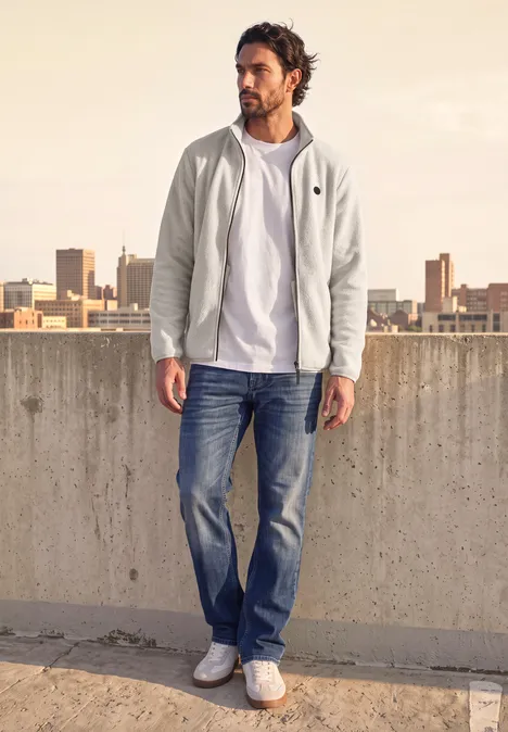Veste en peluche avec col montant et fermeture à zip classic beige Un homme en sweat à capuche blanc, T-shirt, jean et baskets se tient sur un toit-terrasse avec vue urbaine.