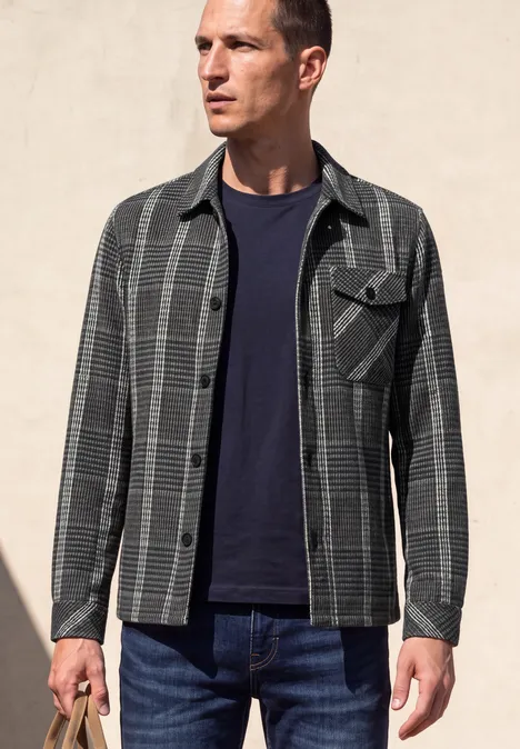 Mann trägt grauschwarze Karo-Hemdjacke über dunkelblauem T-Shirt und Jeans, vor hellem Hintergrund.