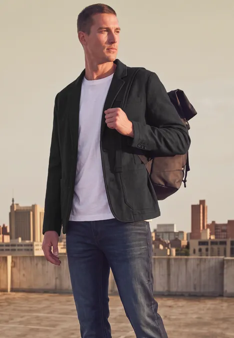 Homme avec sac à dos, veste verte, t-shirt blanc et jeans, debout sur un toit face à la ville.