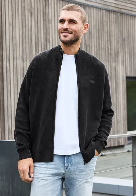 Vest met opstaande kraag en een mix van texturen Black Een glimlachende man in een zwarte jas, wit shirt en spijkerbroek leunt ontspannen tegen een reling.