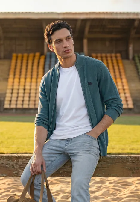 Ein junger Mann in Jeans und Jacke lehnt mit Tasche vor leeren Tribünen auf einem Sportplatz.