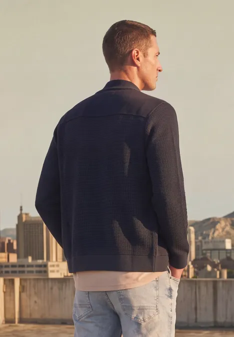 Strickjacke mit Stehkragen und Struktur dark basic blue Mann lehnt auf Dachgeländer, trägt Jacke und Jeans, Blick auf Stadt und Berge im Hintergrund.