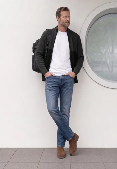 Ein lässig gekleideter Mann lehnt an einer Wand neben rundem Fenster und trägt Jeans, weißes Shirt, Jacke und Rucksack.