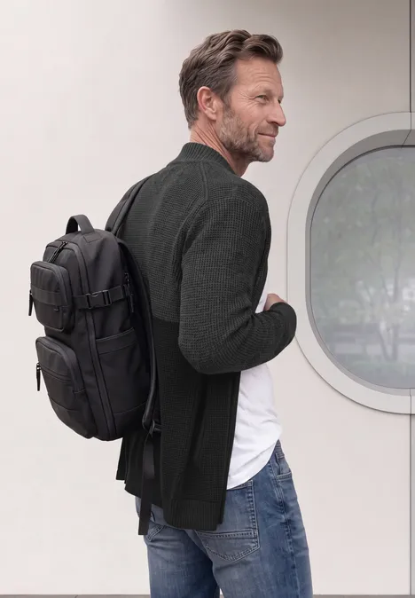 Ein Mann in Jeans und Sakko trägt einen schwarzen Rucksack und sieht seitlich eine Hausfassade mit rundem Fenster.