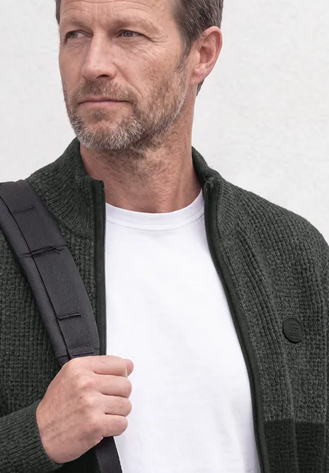 Ein Mann mittleren Alters mit grauem Bart trägt grüne Strickjacke, weißes T-Shirt und Umhängetasche.