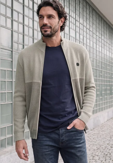 Ein Mann trägt eine beige Strickjacke, ein dunkelblaues T-Shirt und Jeans vor einer Glassteinwand.