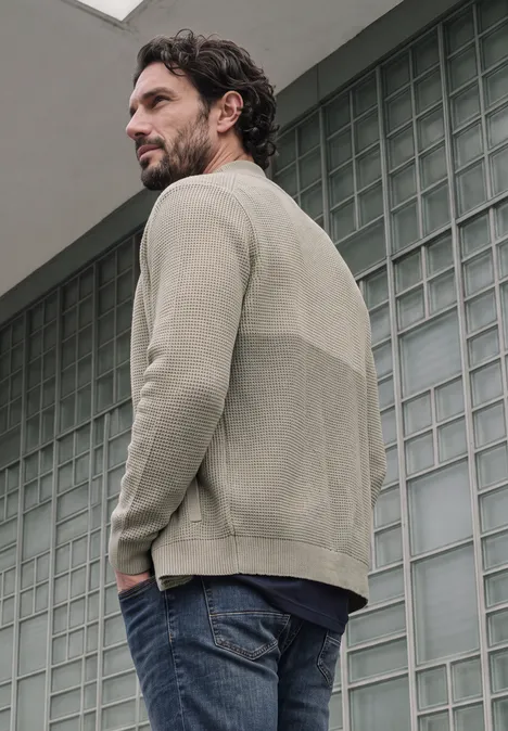 Ein Mann in Jeans und beigem Pullover steht seitlich vor einer Glasbausteinwand, Hände in den Taschen.