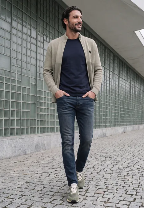 Ein Mann mit Bart in beiger Jacke, dunkelblauem T-Shirt und Jeans geht auf einem Gehweg aus Pflastersteinen.