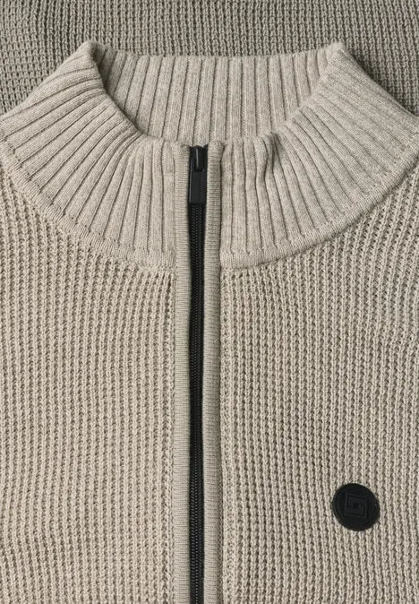 Beige Strickjacke mit hohem Kragen, Reißverschluss mittig und kleinem schwarzen Rundpatch auf der Vorderseite.