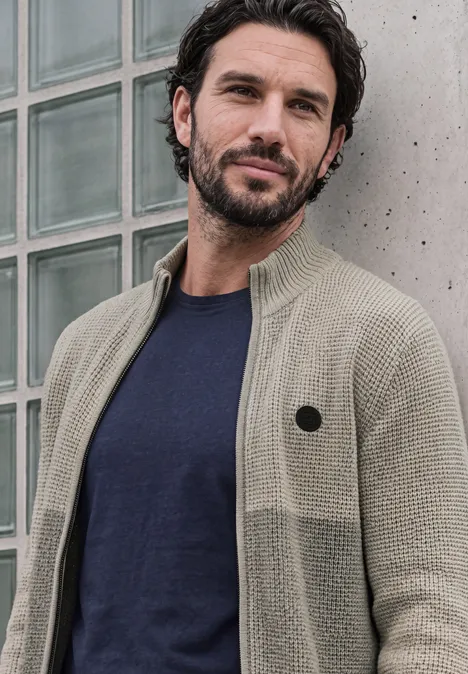 Ein Mann mit dunklen Haaren und Bart trägt einen beigen Strickcardigan vor einer Glaswand.