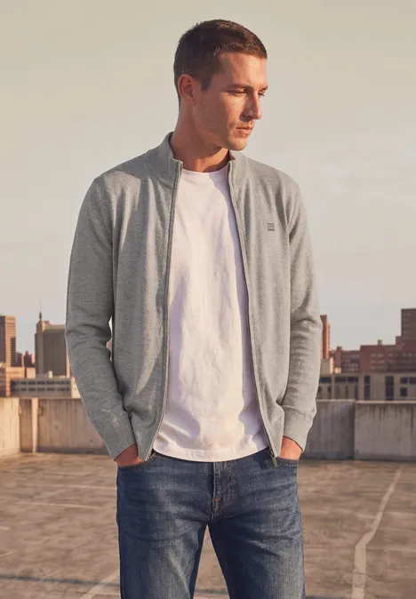 Ein junger Mann in grauem Cardigan, weißem T-Shirt und Jeans steht auf einem Dach und schaut zur Seite.