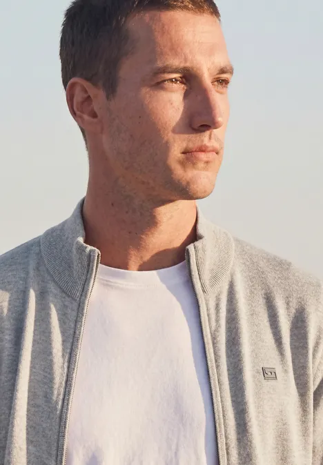 Ein junger Mann steht im natürlichen Licht und trägt ein weißes T-Shirt und eine graue Jacke.
