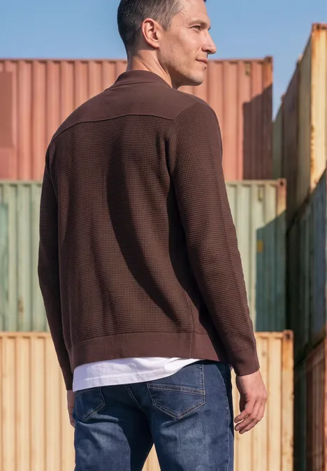 Ein Mann in brauner Jacke und Jeans steht vor bunten Frachtcontainern und blickt weg.