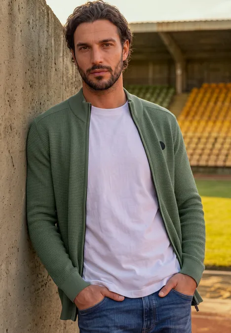 Ein Mann lehnt lässig an einer Betonwand vor Stadionrängen, trägt grüne Jacke und weißes T-Shirt.