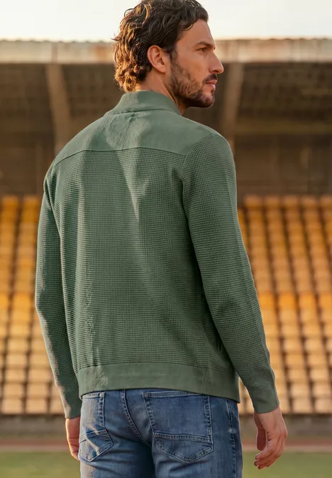 Mann steht auf einem Sportplatz vor leeren Tribünen, trägt grüne Jacke und Jeans.
