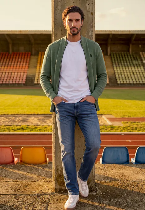 Ein Mann in Jeans, weißem T-Shirt und grüner Jacke steht lässig im Stadion vor Tribünen.