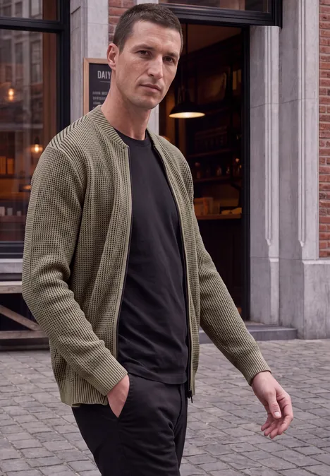 Un homme en gilet vert et t-shirt noir marche devant un café en ville.