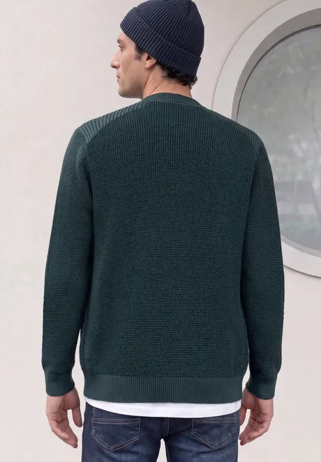 Homme vu de dos, pull vert foncé, sous-vêtement blanc dépassant, bonnet, devant une fenêtre ronde.