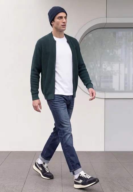 Un homme en cardigan, T-shirt, jeans et bonnet marche devant une façade moderne.